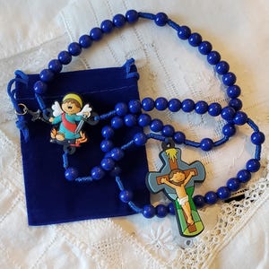 May include: A blue rosary with round beads and a blue drawstring pouch. The rosary features a cross pendant with a depiction of Jesus and an angel pendant. The rosary is displayed on a white, lace-trimmed surface.