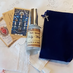 May include: A selection of religious items, featuring a bottle of "Pure Blessed Holy Water", prayer cards, and a navy blue velvet pouch with a silver cross. The items are displayed on a white, lace-trimmed surface.
