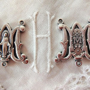May include: Two silver-toned metal charms in the shape of the letter "M". One charm features a depiction of a religious figure, the other has a cross and the letter "M". The charms are on a white, textured background.