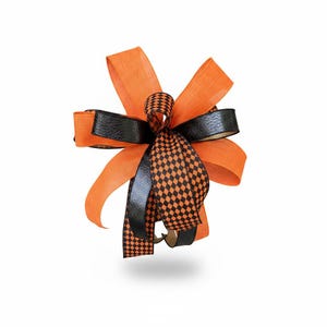 May include: A decorative bow featuring multiple loops made from orange fabric, solid black textured ribbon, and a black and orange checkered pattern ribbon. The bow is arranged with several layers of ribbon extending outward in a starburst shape.