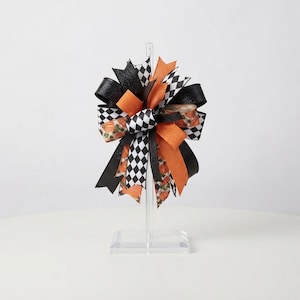 May include: A decorative bow mounted on a clear acrylic stand. The bow features a mix of solid orange ribbons, black glittery ribbons, and ribbons with a black and white checkered pattern or a print of small orange pumpkins on a neutral background.