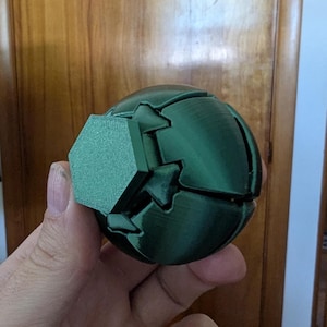 Puede incluir: Un objeto esférico de color verde oscuro impreso en 3D que presenta un panel plano hexagonal texturizado en su exterior. La superficie muestra líneas de capa visibles características de la fabricación aditiva, con paneles geométricos segmentados y entrelazados.