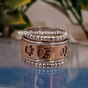 Puede incluir: Un anillo giratorio de plata y oro con huellas de patas grabadas en la banda. El anillo está hecho de plata 925 y tiene una banda giratoria.
