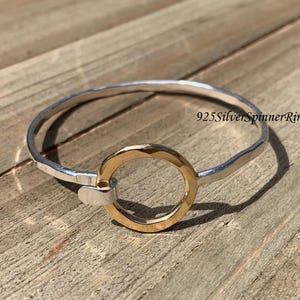 Puede incluir: Un brazalete de plata con acabado martillado y un dije circular de oro. El brazalete está abierto y tiene un diseño simple.