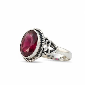 May include: A sterling silver ring featuring a large, oval-cut, deep red gemstone. The gemstone is set in a decorative bezel with a twisted rope design. The ring has a heart-shaped detail on the band and is marked with "925".