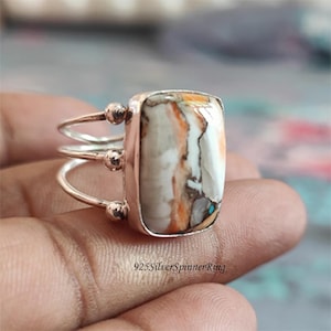 Könnte beinhalten: Ein Silberring mit einem rechteckigen, mehrfarbigen Stein. Der Stein weist weiße, orangefarbene und braune Muster auf. Der Ring hat ein einzigartiges Design mit drei kleinen Silberkugeln auf jeder Seite des Steins. Der Text "925SilverSpinnerRing" ist sichtbar.