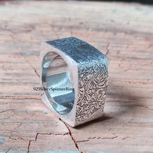 May include: A square silver ring with a floral pattern etched on the surface. The ring has a textured appearance and is set against a wooden background. The text "925SilverSpinnerRing" is visible.