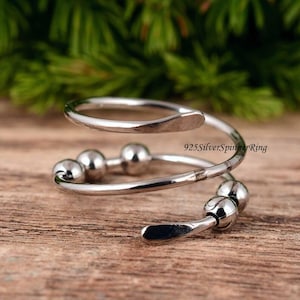 Könnte beinhalten: Ein silberfarbener Spinner-Ring mit Spiraldesign. Der Ring hat kleine, runde Kugeln und ein glattes, gebogenes Band. Der Text "925SilverSpinnerRing" ist auf dem Ring sichtbar.