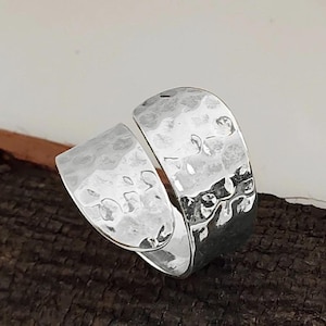 Pode incluir: Um anel prateado com textura martelada. O anel tem um design aberto, com duas bandas curvas e sobrepostas. A superfície do anel reflete a luz, destacando o padrão martelado. O anel é exibido em uma superfície marrom escura.