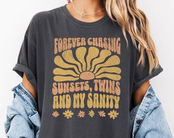 May include: A dark grey t-shirt with a retro-style graphic that says "Forever chasing sunsets, twins and my sanity". The graphic features a stylised sun with a yellow and orange colour scheme and small flowers.