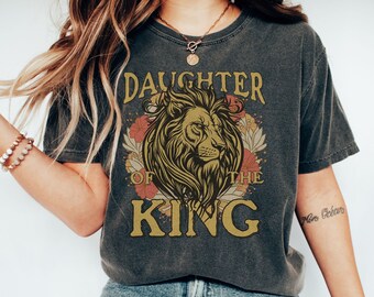 May include: A charcoal gray t-shirt with a vintage-style graphic of a lion with floral accents. The text "Daughter of the King" is printed on the shirt.