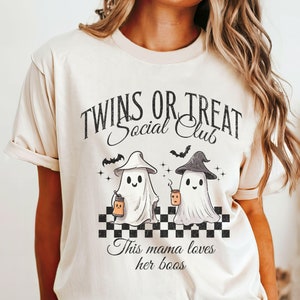 Puede incluir: Una camiseta beige con un gr&aacute;fico de dos fantasmas sosteniendo cubos de dulces. El texto en la camiseta dice "Twins or Treat Social Club" y "This mama loves her boos".