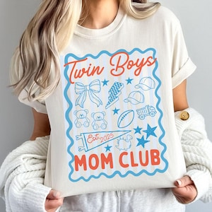 May include: Cream-colored t-shirt with a blue and red graphic that reads "Twin Boys Mom Club Est. 2025." The graphic includes illustrations of a bow, lightning bolt, baseball caps, teddy bears, a football, a truck, and stars.