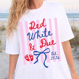 May include: White t-shirt featuring the text "Red, White & Due" in red and blue script, with a blue bow and baby footprints. Pink and white vertical stripes are behind the text. The text "Est. 2026" is also on the shirt.