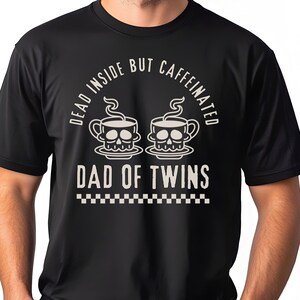Op de afbeelding: Zwart T-shirt met de tekst "DEAD INSIDE BUT CAFFEINATED" boven twee koffiekopjes in de vorm van een schedel en de woorden "DAD OF TWINS" eronder. De onderkant van het shirt heeft een dambordpatroon.