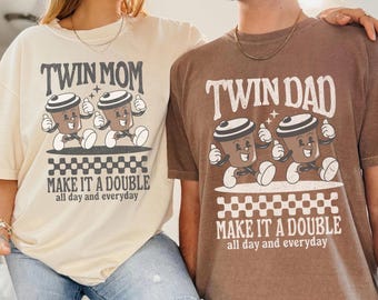 May include: A cream-coloured t-shirt with the text TWIN MOM MAKE IT A DOUBLE all day and everyday and a brown t-shirt with the text TWIN DAD MAKE IT A DOUBLE all day and everyday, both featuring graphic illustrations of two coffee cups.