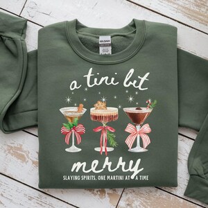 Può includere: Felpa verde oliva con la scritta "a tini bit merry" e tre bicchieri da martini con decorazioni festive. Il testo "Slaying Spirits, One Martini at a Time" è stampato sotto la parola "merry".