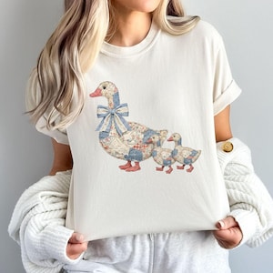 Puede incluir: Camiseta color crema con un diseño de patchwork de una madre pato y dos patitos. El pato tiene un lazo de cuadros azul y pies rojos. Los patitos también tienen detalles de patchwork. La camiseta tiene mangas enrolladas.