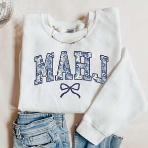 May include: A white sweatshirt featuring the word "MAHJ" in blue floral print lettering and a blue bow graphic. The sweatshirt is styled with blue jeans and a delicate necklace.