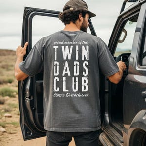 May include: A charcoal gray t-shirt with the text "TWIN DADS CLUB" in white block letters. The shirt also features the phrases "proud member of the" and "Classic Overachiever". The person is standing next to a vehicle.