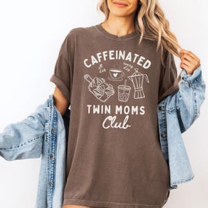 May include: A brown t-shirt with white text that reads "Caffeinated all day every day" and "Twin Moms Club". The shirt features a coffee cup, a glass of iced coffee, and a coffee pot.