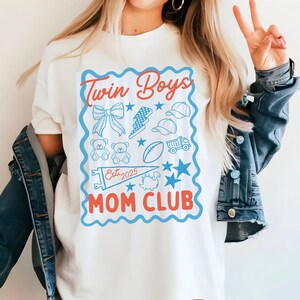 May include: White t-shirt with a blue and red graphic that reads "Twin Boys Mom Club Est. 2025." The graphic includes illustrations of a bow, lightning bolt, baseball caps, teddy bears, a football, a truck, stars, and a dinosaur.