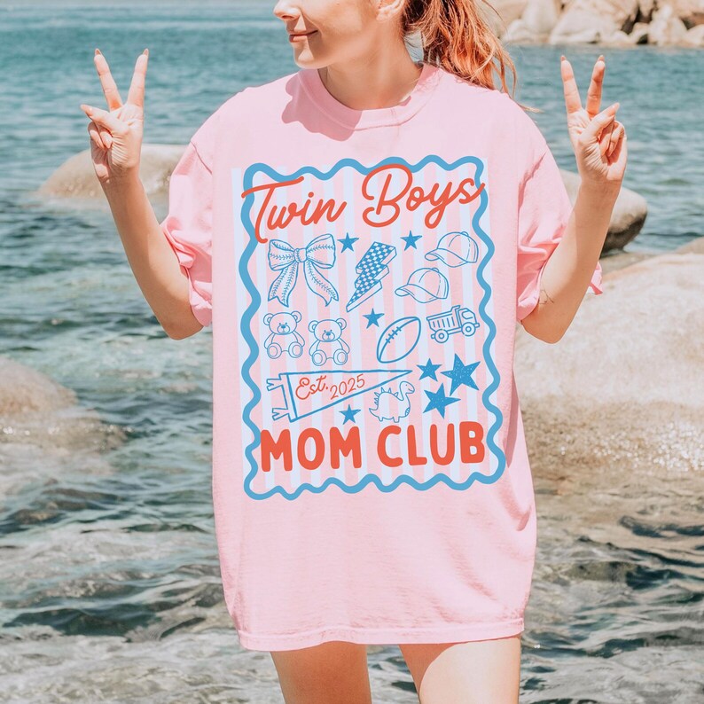 May include: Pink t-shirt with a blue and white graphic that says "Twin Boys Mom Club Est. 2025". The graphic includes a bow, lightning bolt, baseball caps, teddy bears, a football, a truck, stars, and a pennant.