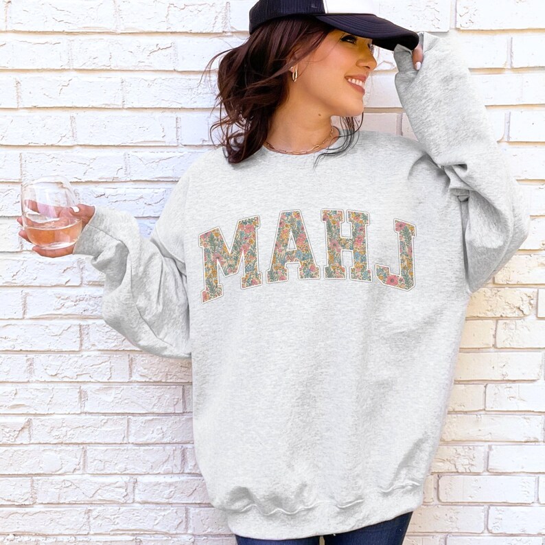 May include: A light gray sweatshirt with the word "MAHJ" in floral print letters. The sweatshirt has long sleeves and a crew neck. The person in the image is wearing a black and white baseball cap.