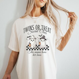 Puede incluir: Una camiseta blanca con un gr&aacute;fico de dos fantasmas sosteniendo tazas. El texto en la camiseta dice "Twins or Treat Social Club" y "This mama loves her boos".