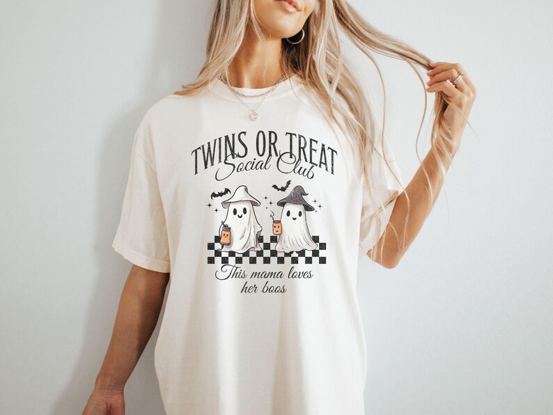 Puede incluir: Una camiseta blanca con un gr&aacute;fico de dos fantasmas sosteniendo tazas. El texto en la camiseta dice "Twins or Treat Social Club" y "This mama loves her boos".