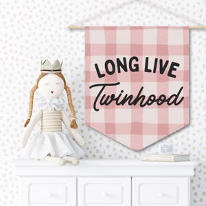 Könnte beinhalten: Ein rosa-weiß kariertes Banner mit dem Text "LONG LIVE Twinhood" hängt an einer Wand. Eine Puppe mit Krone und Zöpfen sitzt auf einer weißen Kommode. Das Banner ist ein dekorativer Artikel für ein Kinderzimmer.