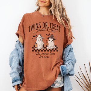 Puede incluir: Una camiseta de color &oacute;xido con un gr&aacute;fico de dos fantasmas sosteniendo tazas. El texto en la camiseta dice "Twins or Treat Social Club" y "This mama loves her boos".