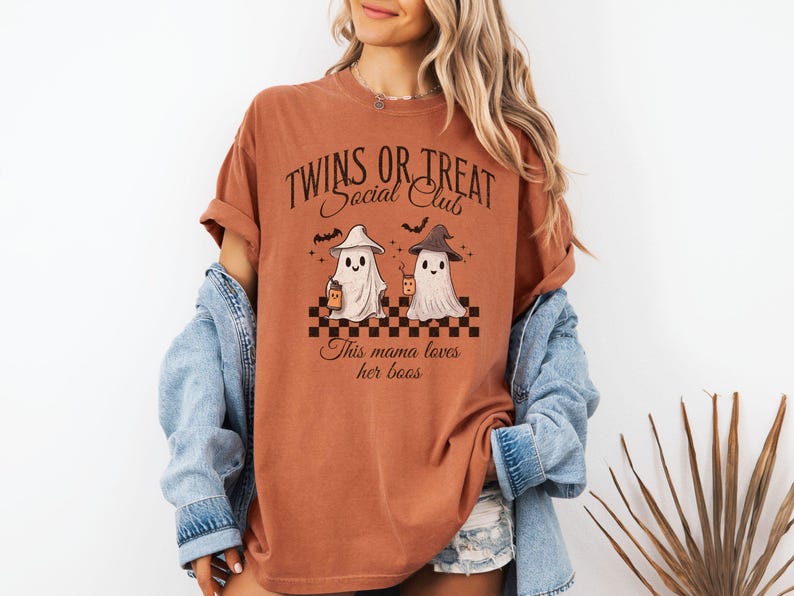 Puede incluir: Una camiseta de color &oacute;xido con un gr&aacute;fico de dos fantasmas sosteniendo tazas. El texto en la camiseta dice "Twins or Treat Social Club" y "This mama loves her boos".