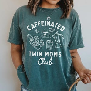 May include: Teal blue t-shirt with a white graphic design that says "caffeinated all day, every day" with images of coffee, tea, and pastries. The text "Twin Moms Club" is below the graphic.
