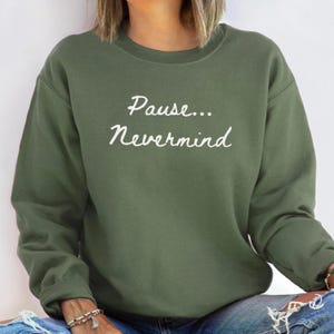 May include: An olive green sweatshirt with the words "Pause... Nevermind" in white script. The crew neck sweatshirt is a solid color and has long sleeves.