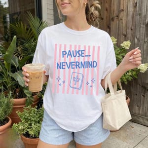 May include: A white short-sleeve t-shirt featuring a graphic with vertical pink stripes, the blue text "PAUSE... NEVERMIND", and a blue outline of a mahjong tile with a character in the center, accented by small blue four-pointed star shapes.