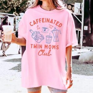 May include: Pink oversized t-shirt with a graphic design that says "Caffeinated all day every day" with a coffee cup, a french press, and a to-go cup. The graphic also says "Twin Moms Club".