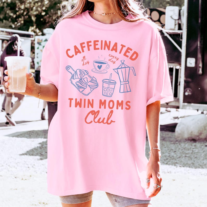 May include: Pink oversized t-shirt with a graphic design that says "Caffeinated all day every day" with a coffee cup, a french press, and a to-go cup. The graphic also says "Twin Moms Club".