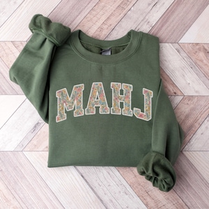 May include: An olive green sweatshirt with the letters "MAHJ" in a floral pattern across the chest. The sweatshirt has long sleeves with ribbed cuffs. The garment is folded on a light brown wooden surface.