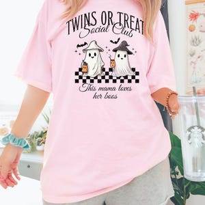 Puede incluir: Camiseta rosa con un gr&aacute;fico de dos fantasmas sosteniendo tazas y el texto "Twins or Treat Social Club" y "This mama loves her boos".