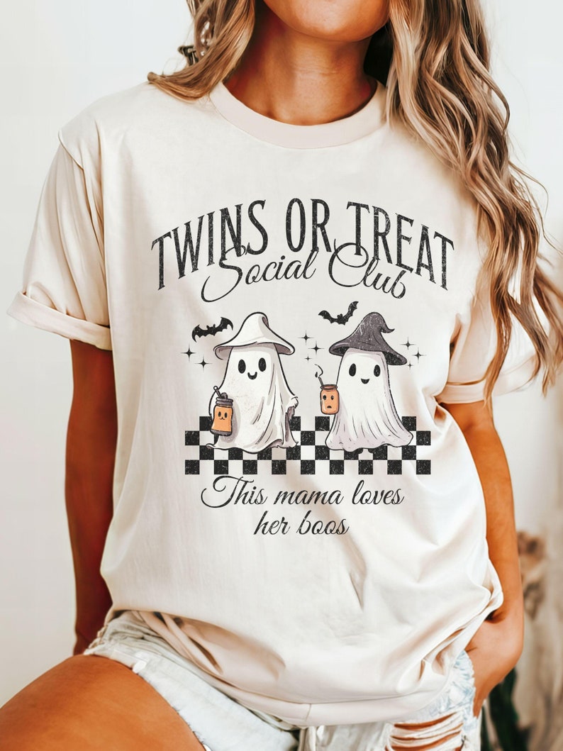 Puede incluir: Una camiseta beige con un gr&aacute;fico de dos fantasmas sosteniendo cubos de dulces. El texto en la camiseta dice "Twins or Treat Social Club" y "This mama loves her boos".