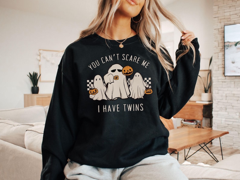 May include: Black sweatshirt with a white graphic of three ghosts holding jack-o-lanterns. The text on the sweatshirt reads 'You can't scare me I have twins'.