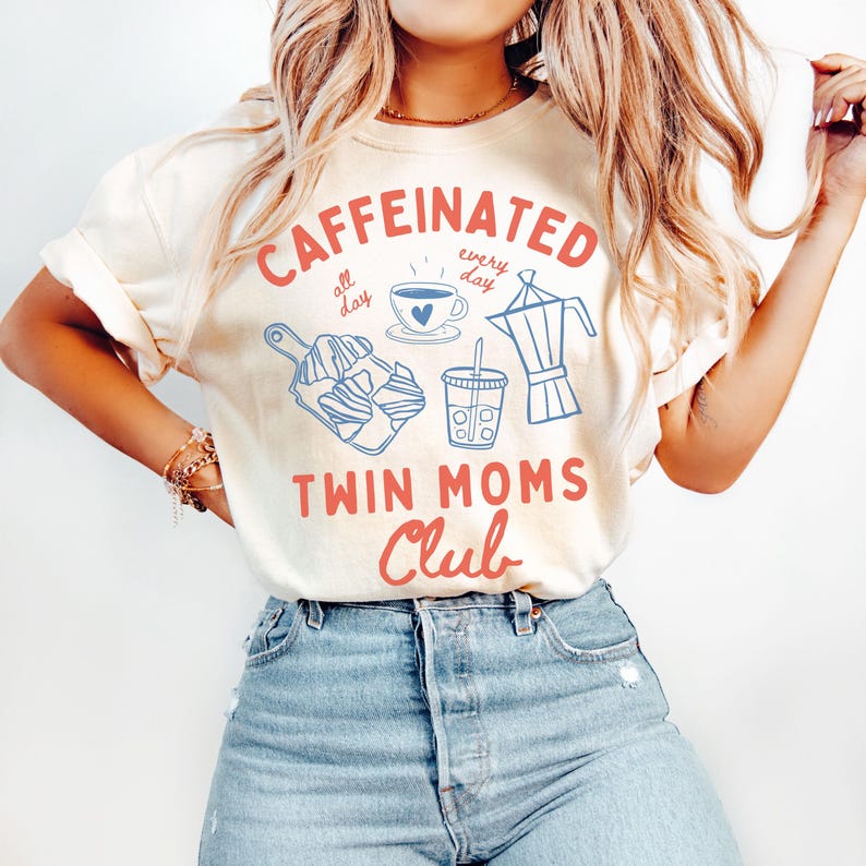 May include: A light yellow t-shirt with the text "CAFFEINATED all day every day" in blue and red. The shirt also features a coffee cup, a coffee pot, and a pastry. The text "TWIN MOMS CLUB" is in red.