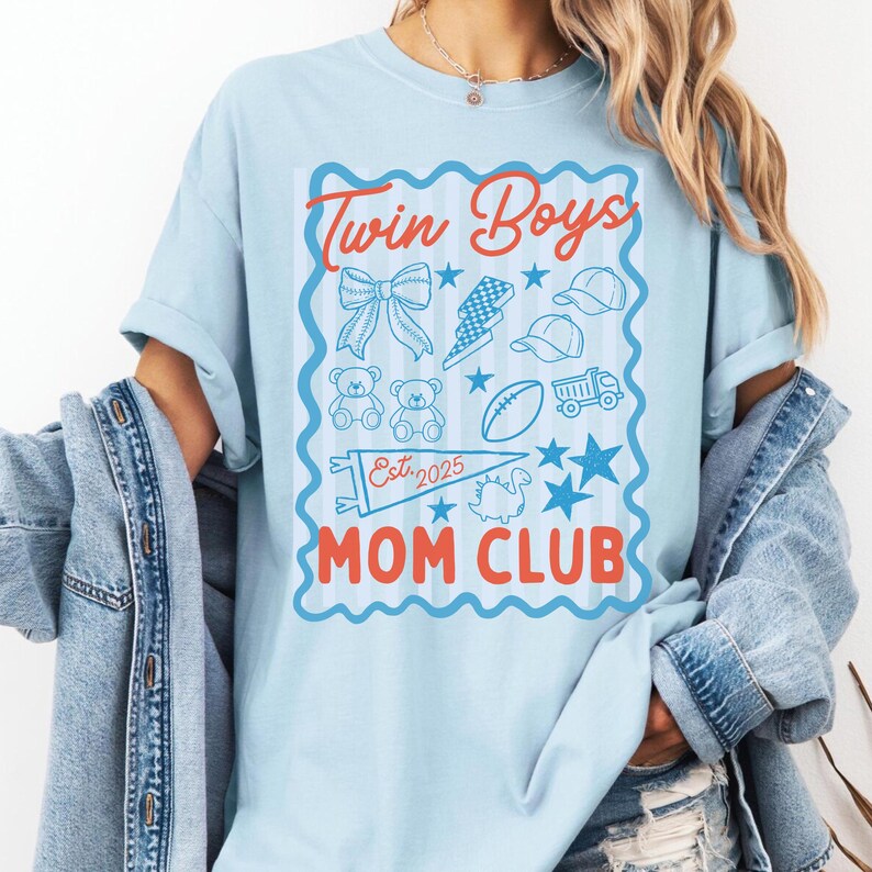 May include: Light blue t-shirt with a graphic that reads "Twin Boys Mom Club" in red and blue. The graphic includes illustrations of a bow, lightning bolt, baseball caps, teddy bears, a football, a truck, stars, and a dinosaur. The text "Est. 2025" is also included.