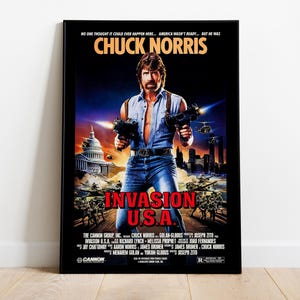 May include: A framed movie poster for "Invasion U.S.A." featuring Chuck Norris. The poster has a black border and a dark background with the title in bold yellow and red text. Norris is holding two guns, with military vehicles and a city skyline in the background.