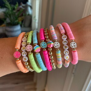 Puede incluir: Un conjunto de pulseras de cuentas de colores con cuentas doradas y dijes de caritas sonrientes. Las pulseras deletrean "TEACH" en letras blancas con letras doradas.