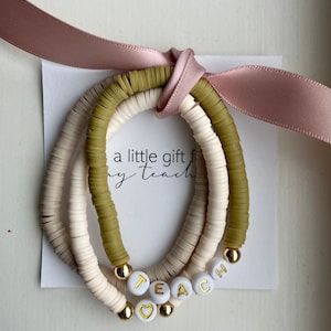 Może przedstawiać: Zestaw trzech bransoletek z białymi i zielonymi koralikami. Bransoletki są ułożone jedna na drugiej i mają złote koraliki z literami "TEACH" i zawieszką w kształcie serca. Bransoletki są związane różową wstążką i kartką z napisem "mały prezent dla mojego nauczyciela".