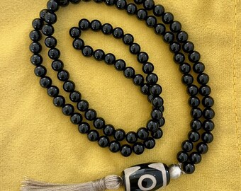 May include: Black beaded necklace with a decorative pendant featuring a white and black design. The necklace has a tassel at the end. The beads are round and shiny. The background is yellow.
