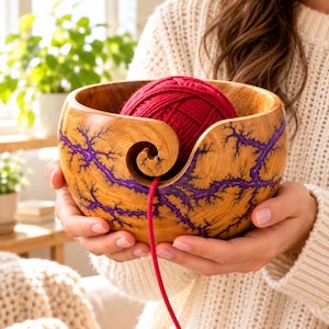 K&ouml;nnte beinhalten: Eine runde Garnschale aus Holz mit nat&uuml;rlicher hellbrauner Maserung und einem tiefvioletten Fraktalmuster. Die Schale hat einen spiralf&ouml;rmigen Ausschnitt an der Vorderseite zur Garnf&uuml;hrung und enth&auml;lt ein rotes Garnkn&auml;uel.