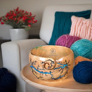 K&ouml;nnte beinhalten: Eine runde Garnschale aus Holz mit nat&uuml;rlicher Holzmaserung und komplizierten, verzweigten Fraktalmustern, die mit leuchtend blauem Harz gef&uuml;llt sind. Die Schale hat einen spiralf&ouml;rmigen Ausschnitt an der Seite f&uuml;r Garn und ein kleines kreisf&ouml;rmiges Logo im Inneren.